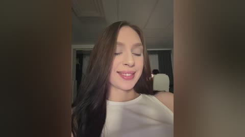 Video of a young woman with fair skin and long dark hair, wearing a white sleeveless top, smiling and sticking her tongue out playfully. Background shows a dimly lit room with a TV and a person in the distance.