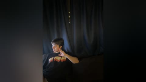 Video of a young Asian man with short black hair, wearing a black graphic T-shirt, sitting in a dimly lit room with dark curtains and a glowing string of fairy lights.