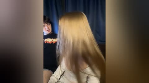 Video of a young woman with long, blonde hair, wearing a white robe, looking at a man with dark hair in a black t-shirt in a dimly-lit room with blue curtains.