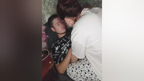 Video of a man in a black t-shirt, eyes closed, lying on a bed, with a woman in a white top and patterned skirt leaning over him, tenderly kissing his cheek.
