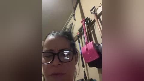 A blurry video of a person with glasses, dark hair, and a neutral expression, standing in a dimly lit room with tools on the wall.