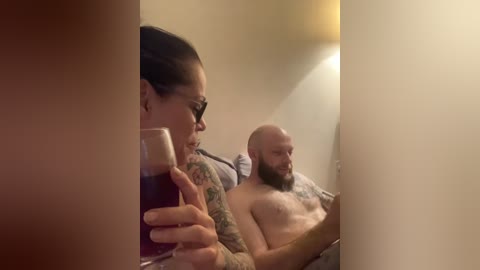 A video shows a tattooed woman holding a beer, seated next to a bald, shirtless man with a beard, both on a bed in a dimly lit room.