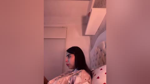 A video of a young woman with long black hair, wearing a patterned robe, lying on a bed with a heart-patterned pillow in a dimly lit room.