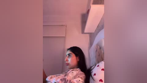 Video of a young woman with long black hair, wearing pajamas with a cartoon character pattern, lying on a bed with a white pillow. The room has a beige wall and a small shelf with a lamp.