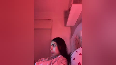 Video of a young woman with long black hair, wearing a pink shirt with hearts, lying on a bed with a heart-patterned pillow, bathed in red light.