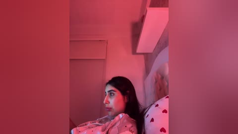 Video of a young woman with long black hair, wearing a polka-dot nightgown, lying in bed with a red light casting a dramatic, eerie ambiance.