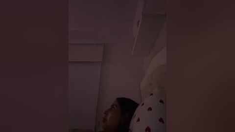 A dimly lit video shows a young girl with brown skin and long dark hair, lying on a bed with a white pillowcase adorned with red hearts. The room is modestly furnished with a white wall and a small window.