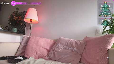 A cozy, festive living room scene with a beige sofa adorned with pink pillows, a lit Christmas tree, a red lamp, and a white TV in the background.