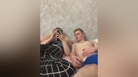Video of a blonde woman in a black and white patterned dress, sitting on a bed next to a shirtless man in blue boxer shorts, both looking at a phone.