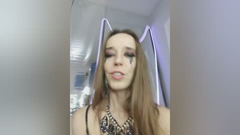 A video of a woman with long, straight brown hair, wearing a black lace top, and adorned with cat ears. She has makeup with bold pink eyeshadow and a small black tear on her right cheek. Background shows a modern, minimalist room with white walls and a metal ceiling.