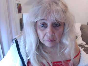 Video of an elderly woman with long, white, curly hair, fair skin, and a serious expression. She wears a red lace bra and a white, textured cardigan. Background shows a bed with white sheets and a door.