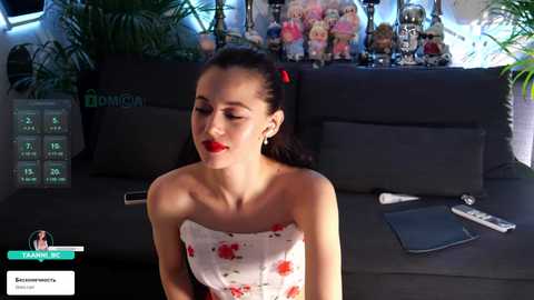 Video of a woman with dark hair in a ponytail, wearing a strapless floral dress, sitting on a black sofa in a room with a TV screen displaying game statistics.