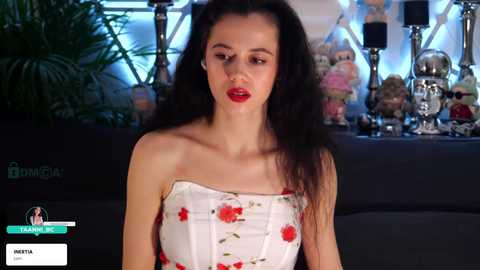 Video of a slender, dark-haired woman in a strapless white dress with red floral pattern, seated indoors. Background includes a dark couch, potted plants, and decorative items.