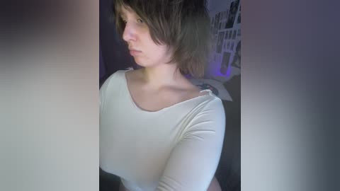 A video of a young woman with short, tousled brown hair, wearing a white, long-sleeved top, looking pensive in a dimly lit room with a blurred, dark background.