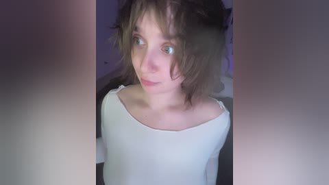 Video of a young woman with messy, shoulder-length brown hair and green eyes, wearing a white, off-the-shoulder top, indoors with blurred background.