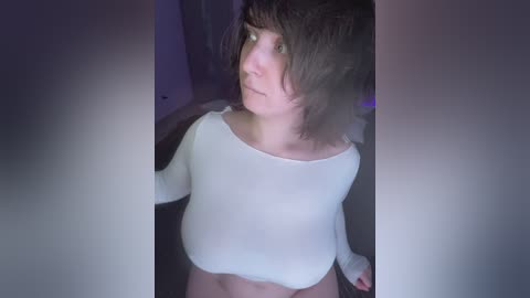 A video of a young woman with shoulder-length brown hair, wearing a white crop top, standing indoors. The background is dimly lit, and her expression appears contemplative.