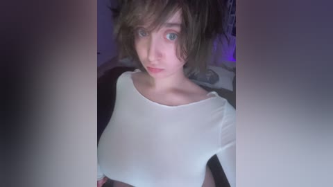 A video of a fair-skinned woman with short brown hair in pigtails, wearing a sheer white top, standing indoors with a blurred background.