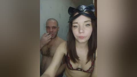 Video of a young woman with fair skin, brown hair, and a black cat headband, wearing a patterned bra, standing next to a bald, muscular, bearded man in a room with beige walls.