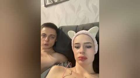 Video of a young, light-skinned woman with long black hair, wearing a pink cat headband, lying next to a shirtless man on a gray couch, in a dimly lit room with white wallpaper.