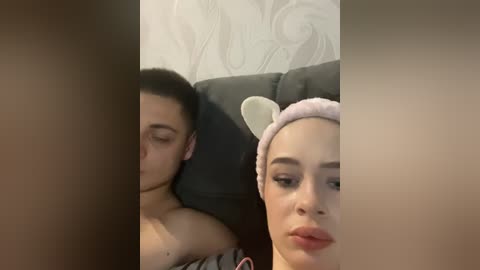 Video of a dark-haired woman with a cat ear headband sleeping on a gray couch, next to a man with short hair. The background features a white patterned wallpaper.