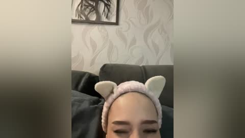 A video of a young woman with fair skin, wearing a light pink headband with cat ears and a matching robe, lounging on a dark grey couch in a softly lit room with floral wallpaper.