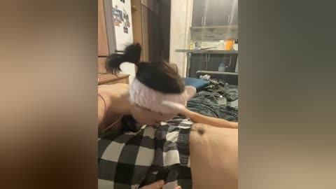 Video of a young Asian woman with black hair in a ponytail wearing a pink headband, lying on a bed with a black and white checkered blanket. The background shows a messy room with a wooden dresser, a mirror, and a window.