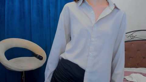Video of a person wearing a white button-down shirt and black skirt, standing in a bedroom with a blue curtain and beige chair in the background.