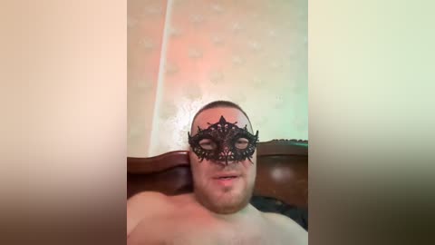 A close-up video of a man wearing an ornate, black masquerade mask with intricate patterns, set against a blurred background with a light-colored wall and a greenish tint.