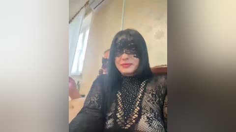 Video of a person wearing a black lace mask and black lace dress with a snake pattern, sitting indoors with a white wall and window in the background.