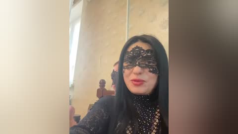 Video of a woman with long black hair, wearing a black lace masquerade mask and a black sequined dress, standing in front of a mirror with a vintage wallpaper background.