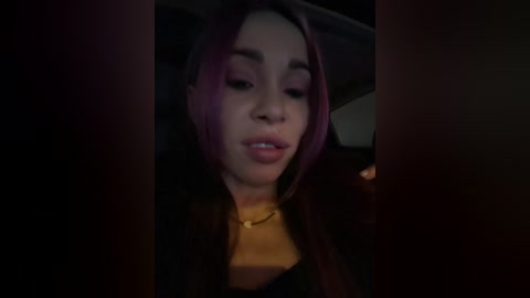 A dimly-lit video shows a young woman with long, dark hair and light skin, wearing a black top and gold necklace, sitting in a car. Her expression is neutral.