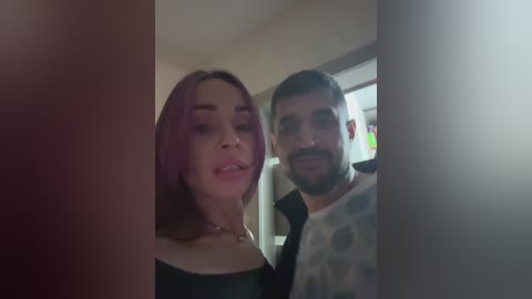 Video of a man with a beard and a woman with long, purple hair, both smiling, standing indoors. The man wears a white shirt with polka dots, and the woman has a black top.