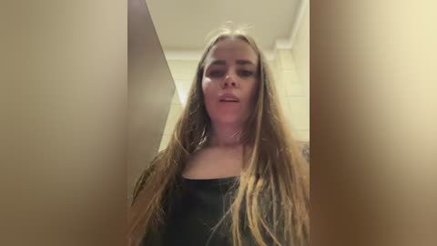 Video of a young, fair-skinned woman with long, light brown hair, wearing a black top, looking directly at the camera. The background is blurred, showing a beige wall and a ceiling.