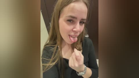 Video of a young Caucasian woman with long brown hair, wearing a black shirt, sticking her tongue out and making a fist in a playful, cheeky pose.
