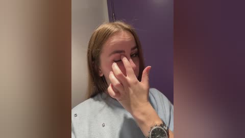 Video of a young woman with light skin, straight brown hair, and a light blue smock, covering her eyes with her right hand, wearing a silver watch on her left wrist.