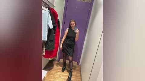 Video of a fair-skinned woman with blonde hair in a black sleeveless mini dress, black tights, and black shoes, taking a mirror selfie in a brightly lit clothing store with purple doors and wooden floors.