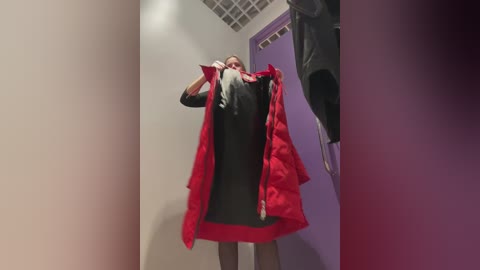 A video of a person in a red and black dress, partially removed, standing in a dimly lit, beige-walled room with a metal grid ceiling. The image is slightly blurred.