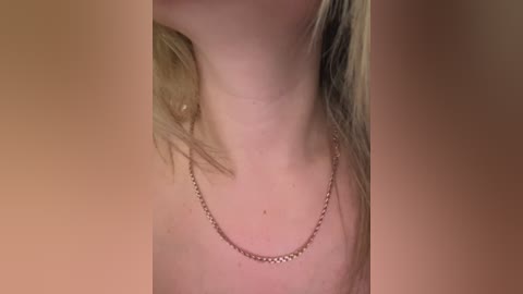 Video of a woman with fair skin, wearing a delicate gold necklace, her blonde hair partially visible, against a blurred, warm-toned background.