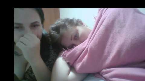 A video of a young woman with curly brown hair, wearing a pink shirt, lying on a bed, partially obscured by a man with a black shirt.