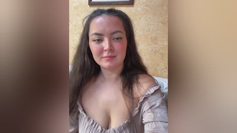 A video of a young woman with long, dark hair, light skin, and a curvy physique, wearing a low-cut, beige blouse, sitting in a room with a textured, light-colored wall and a framed picture in the background.