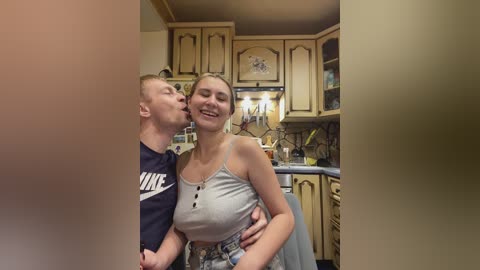 Video of a couple in a cozy, beige-tiled kitchen; man in a dark shirt, woman in a tank top and shorts, both smiling and kissing.