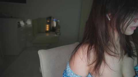 A video of a young woman with long, dark hair, wearing a blue off-shoulder dress, sitting on a beige chair. The background features a dimly lit room with a TV and furniture.