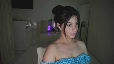 Video of a young woman with dark hair tied in a bun, wearing a blue off-shoulder top, seated in a dimly lit, modern room with a purple lamp and white furniture.