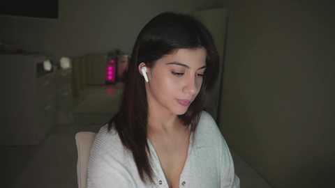 Video of a young woman with straight, dark hair, wearing a light-colored, button-down cardigan, listening to music with white earbuds, in a dimly lit, modern room with a glass table and pink objects in the background.