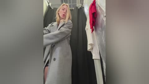 Video of a blonde woman in a gray coat, standing in a changing room with a black curtain, white and red clothes hanging nearby.