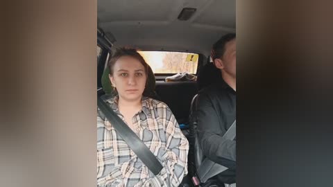 Video of a young woman with shoulder-length brown hair, wearing a plaid shirt, seated in a car with a black seatbelt. A man in a black jacket is partially visible in the background.