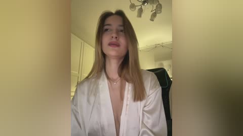 Video of a young woman with light skin and straight, shoulder-length brown hair, wearing a white robe, standing in a dimly lit room with cream-colored walls and a black chair in the background.