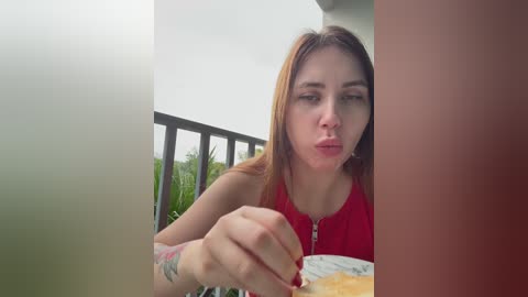 Video of a young woman with light skin, long brown hair, wearing a red tank top, sitting at a table on a balcony, eating a sandwich, with green foliage and a gray sky in the background.