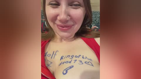 Video of a smiling, fair-skinned woman with brown hair, wearing a red top, with \"Holl king of the daddies 7/3\" tattooed on her chest. Background is blurred.