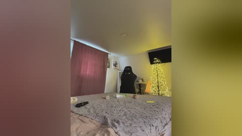 Video of a dimly lit bedroom with a large bed, grey comforter, red curtains, a black gaming chair, a small Christmas tree, and a TV on the wall.